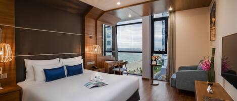A modern hotel room with a large bed, a desk, a chair, and a balcony with a view of the sea.