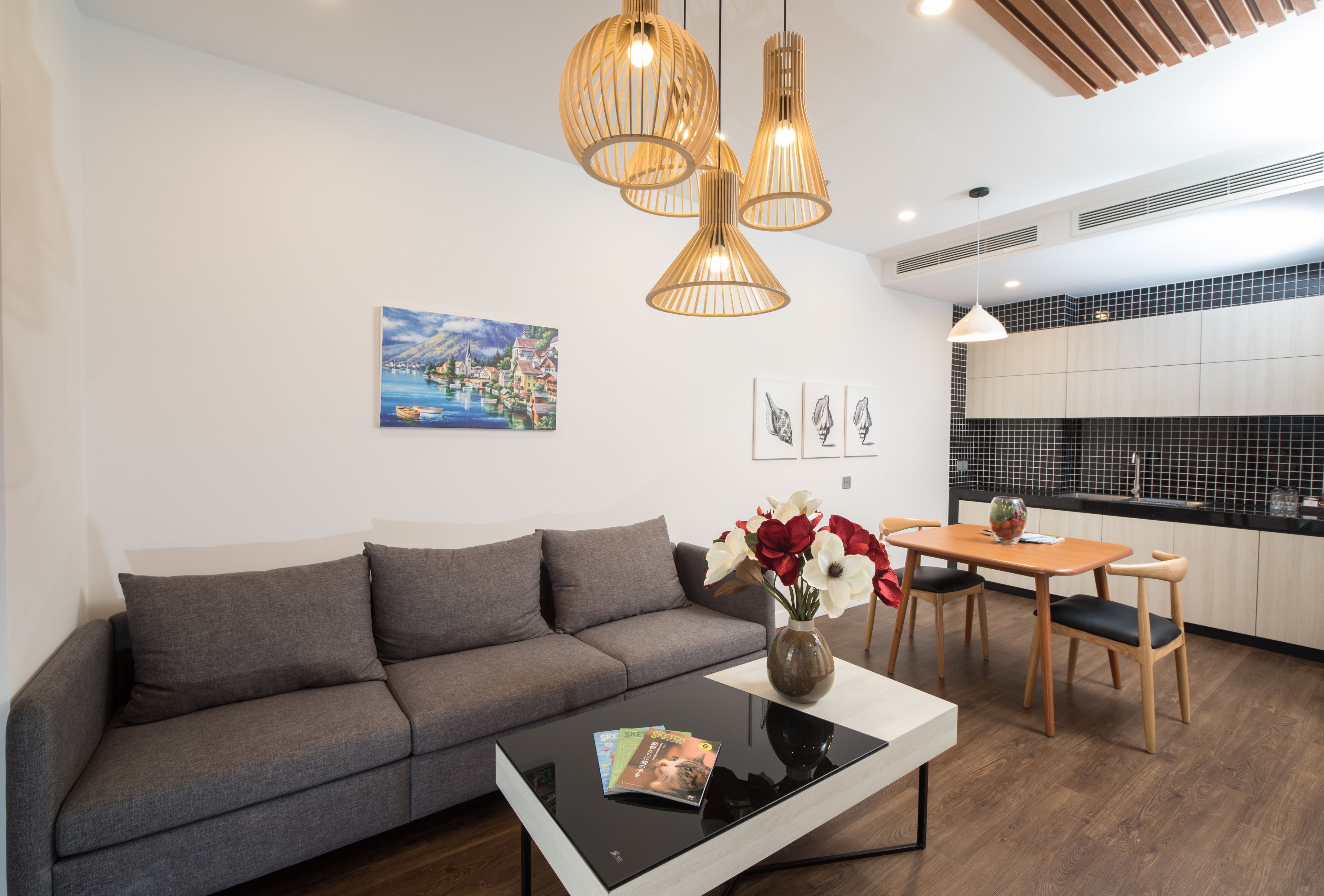 Un salon moderne avec un canapé gris, une table basse noire et un coin repas comprenant des chaises et une table en bois. Trois lampes suspendues sont placées au-dessus du canapé et un tableau orne le mur.