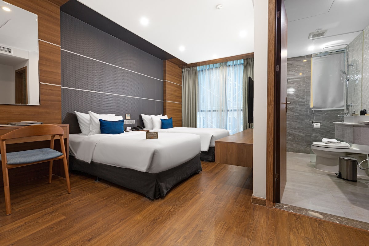 A modern hotel room with two beds, a wooden chair, a desk, and a bathroom with a shower and toilet.