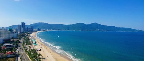 Un paysage urbain côtier avec une vaste plage, une mer bleue et limpide, et une chaîne de montagnes en arrière-plan.
