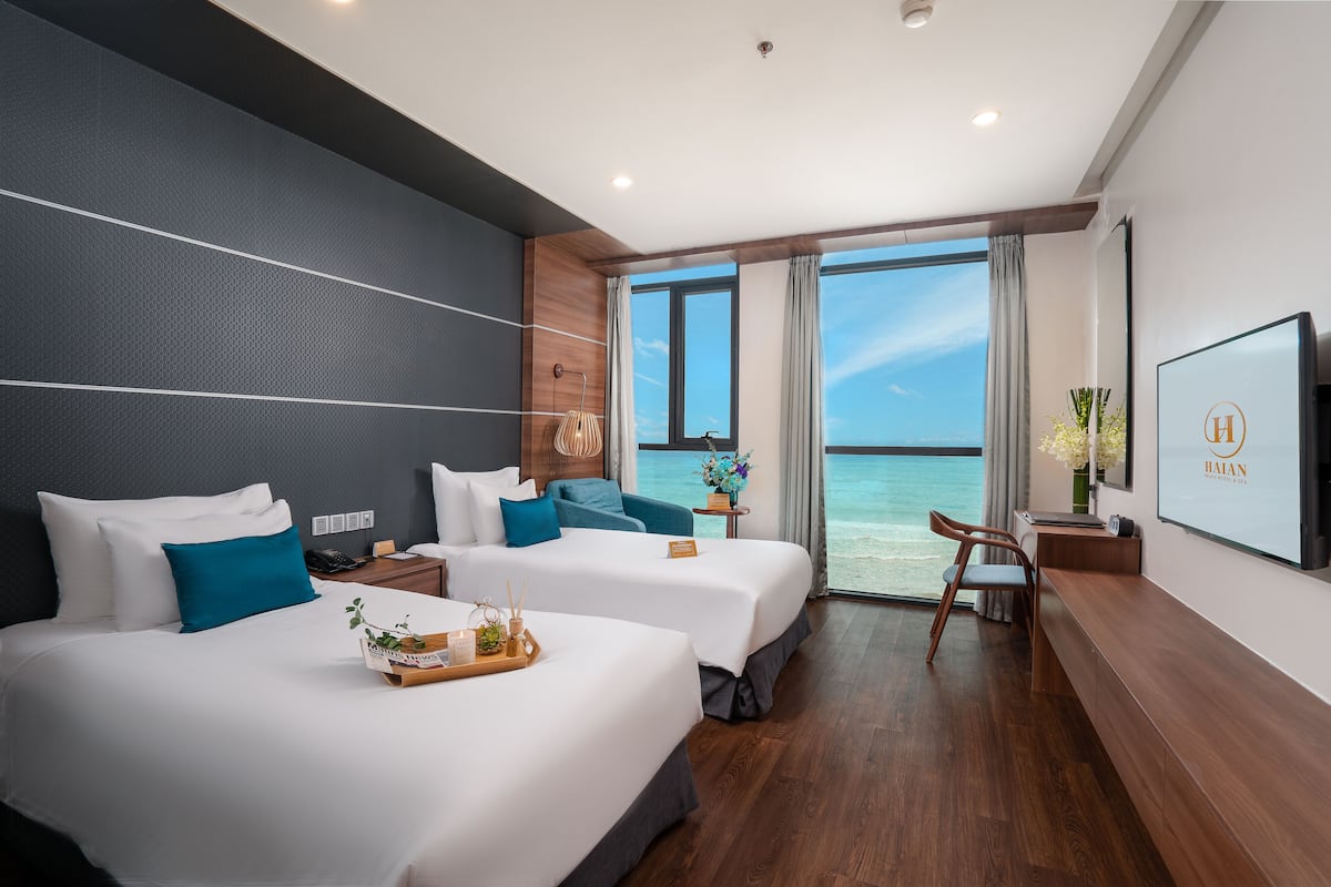 A hotel room with two beds, a desk, a chair, a TV, and a large window with a view of the ocean.