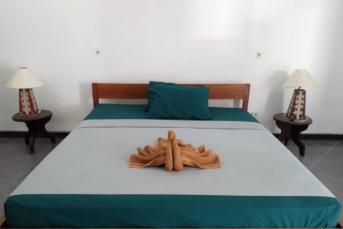 A neatly made bed with a green bedspread and a brown towel arranged in the center. There are two nightstands with lamps on either side of the bed.