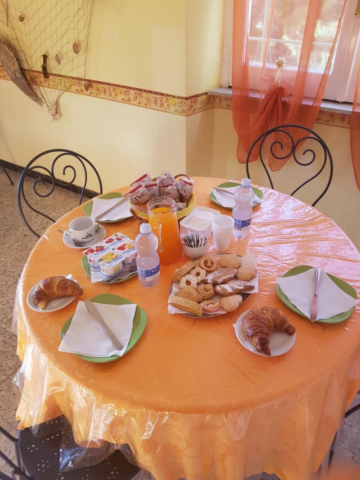 A round table with a plastic tablecloth, covered with plates, napkins, and various food items including pastries and a bottle of orange juice.