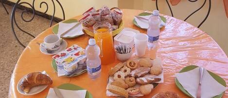 A round table with a plastic tablecloth, covered with plates, napkins, and various food items including pastries and a bottle of orange juice.