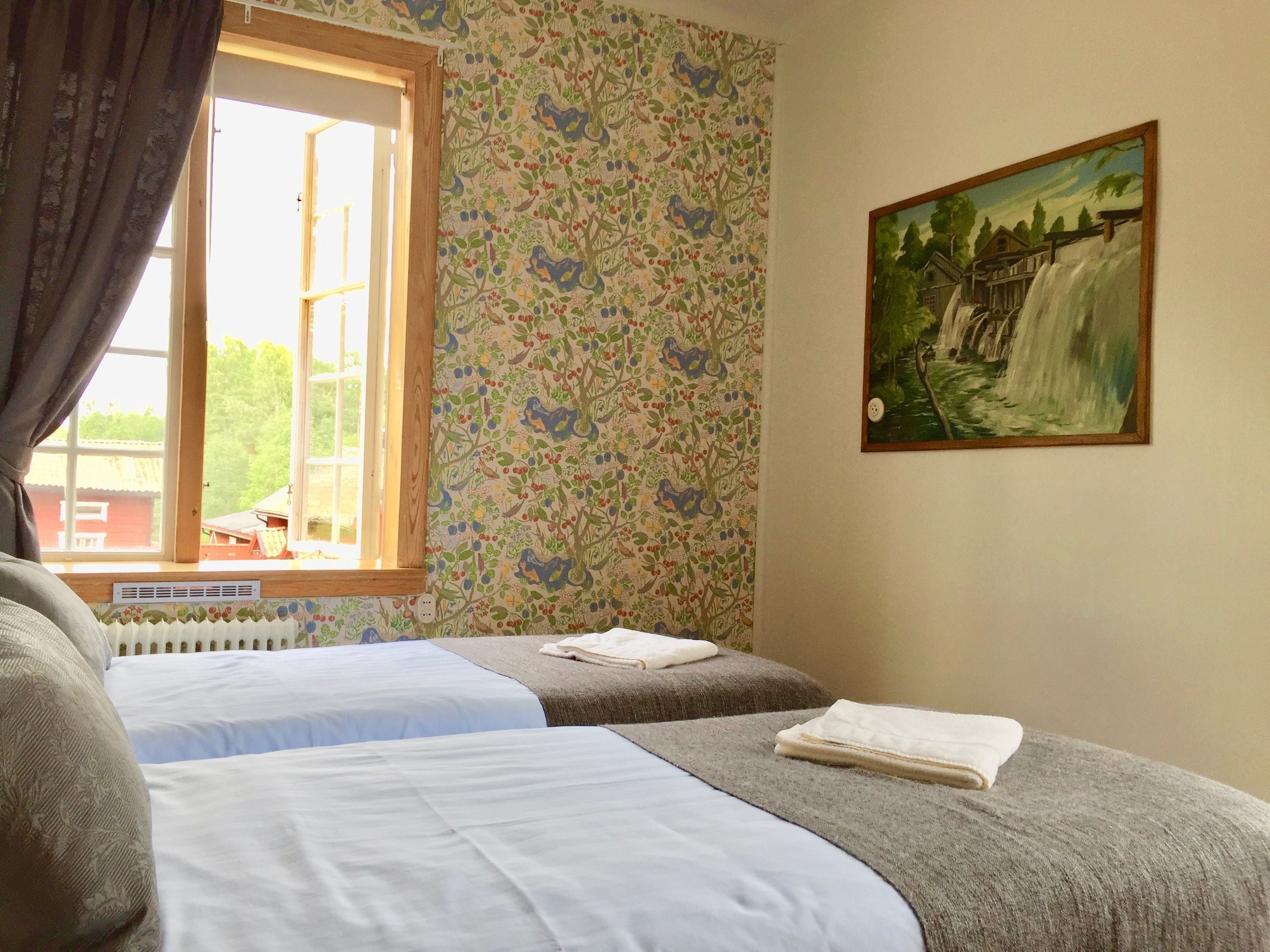 A room with two beds, a window with curtains, a radiator, and a painting on the wall.