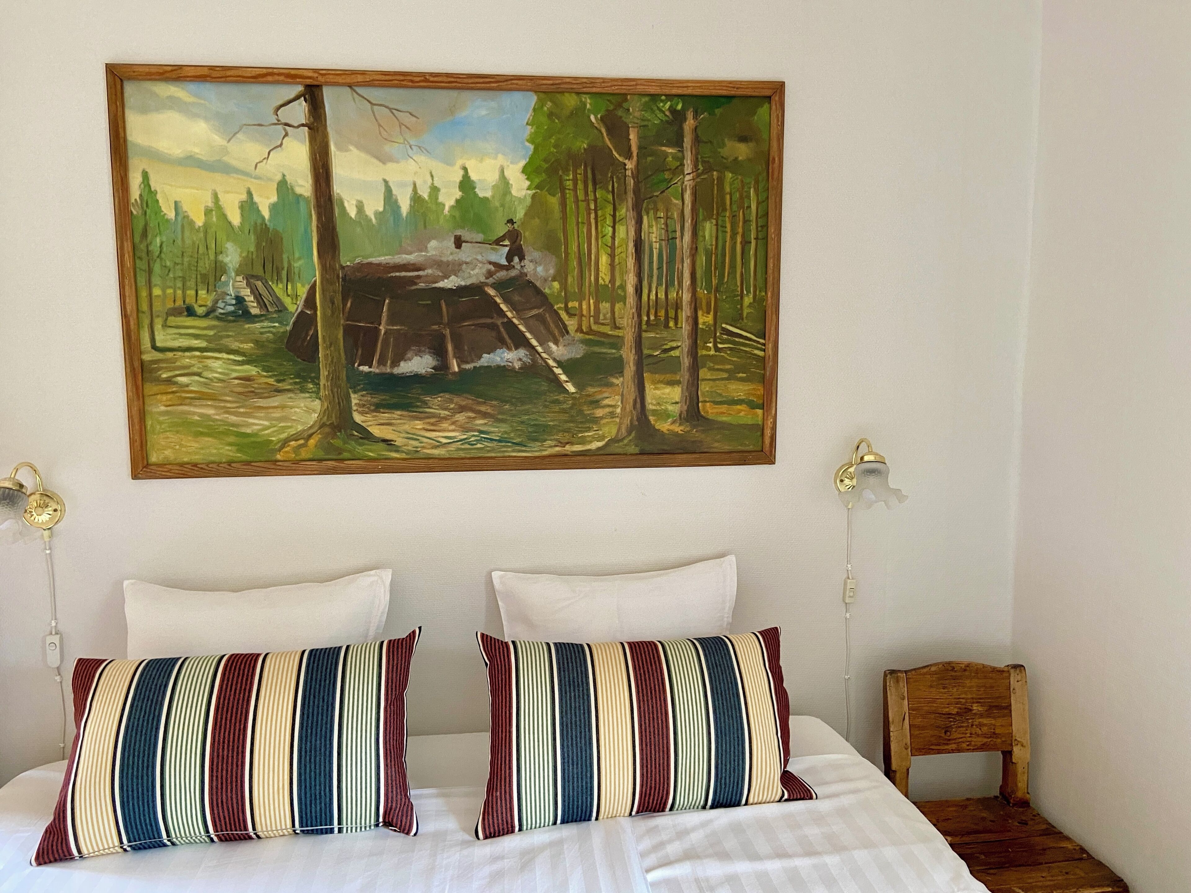 A bedroom with a bed, two striped pillows, a painting of a forest scene, and a small wooden chair.