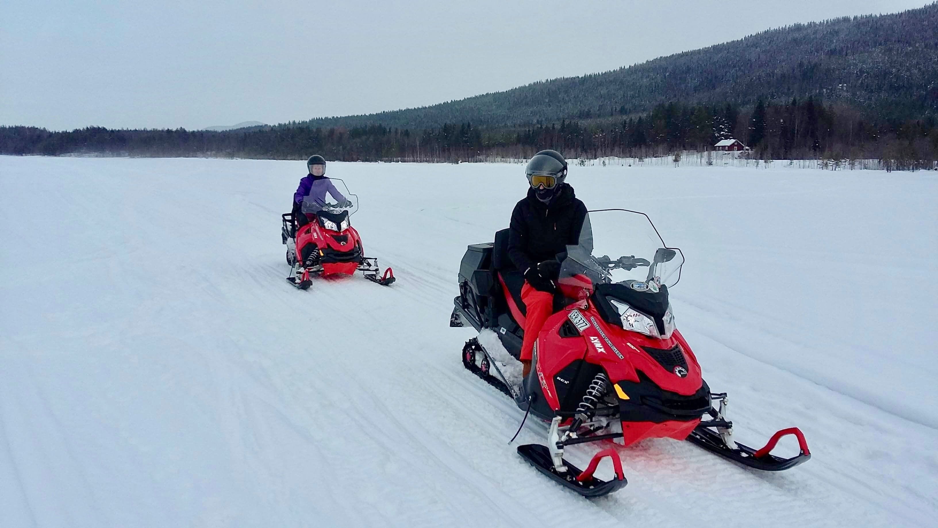 Two snowmobiles on a snowy field with a forested hill in the background.