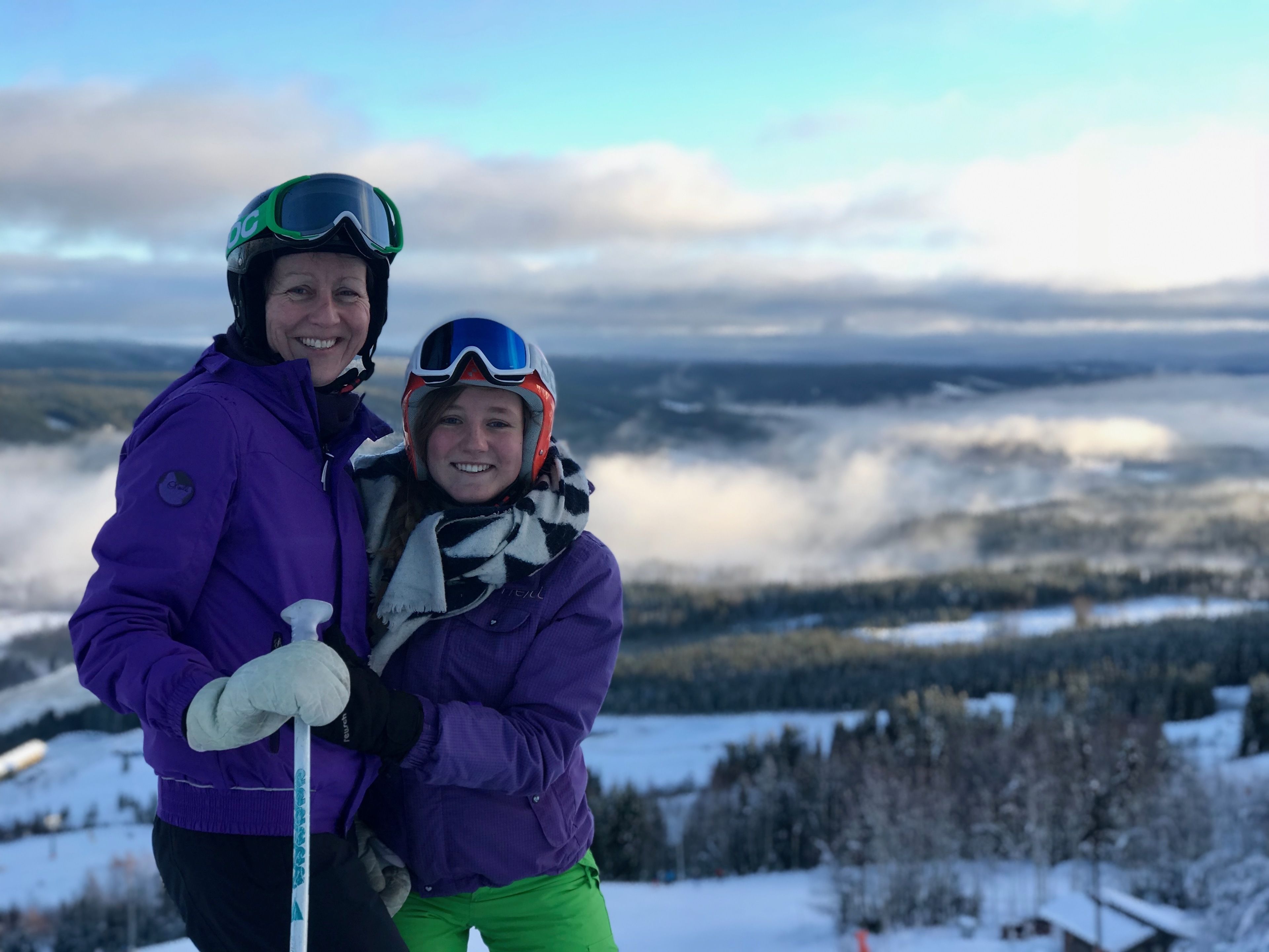 Two people in purple jackets and helmets, standing on a snowy mountain with a view of a forested valley.