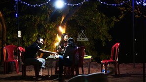 Trois personnes sont assises à une table à l’extérieur, de nuit, sous une guirlande lumineuse.