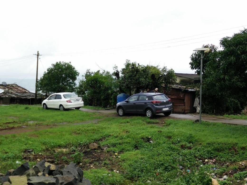 A rural setting with two cars parked on a dirt road, surrounded by greenery and a few buildings.