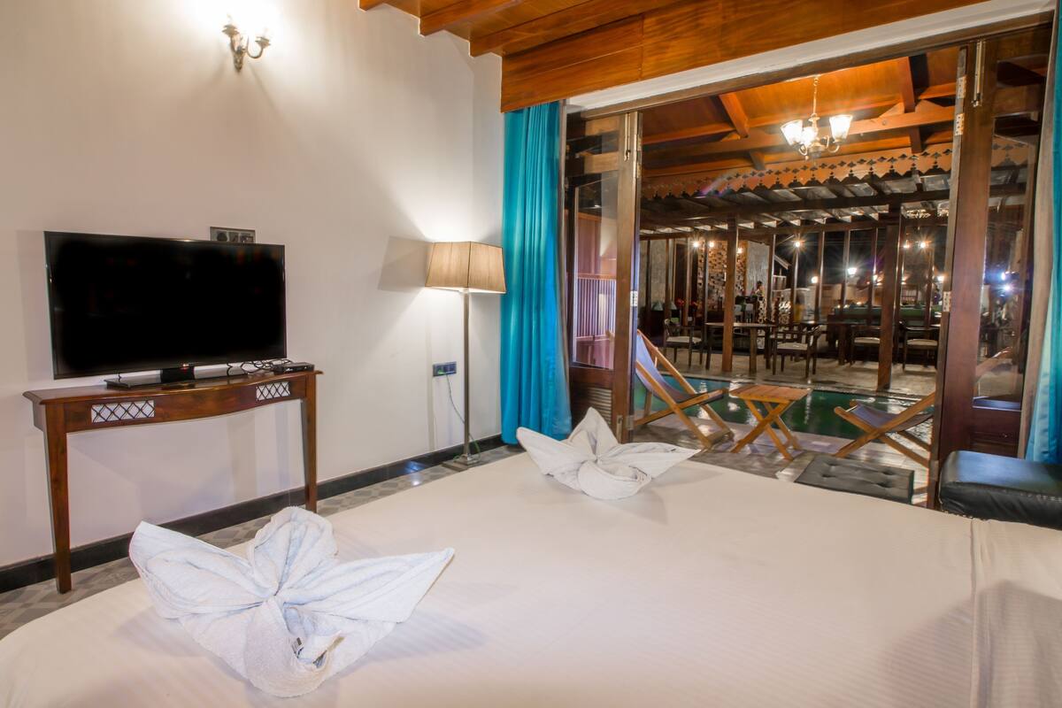 A hotel room with a bed, a TV, a table, and a view of an outdoor pool area.