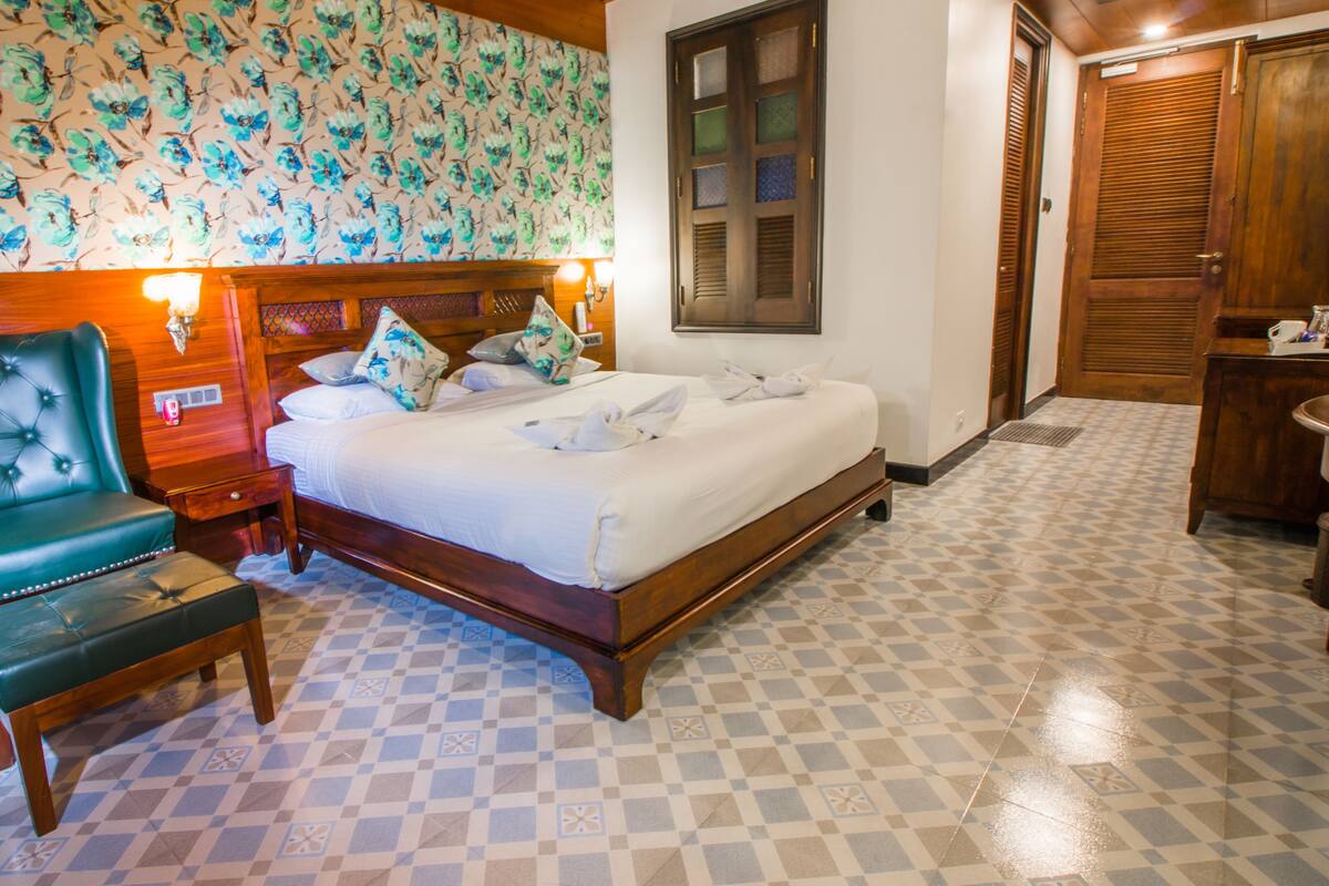 A hotel room with a bed, a green upholstered chair, a wooden headboard, a patterned wallpaper, and a tiled floor.