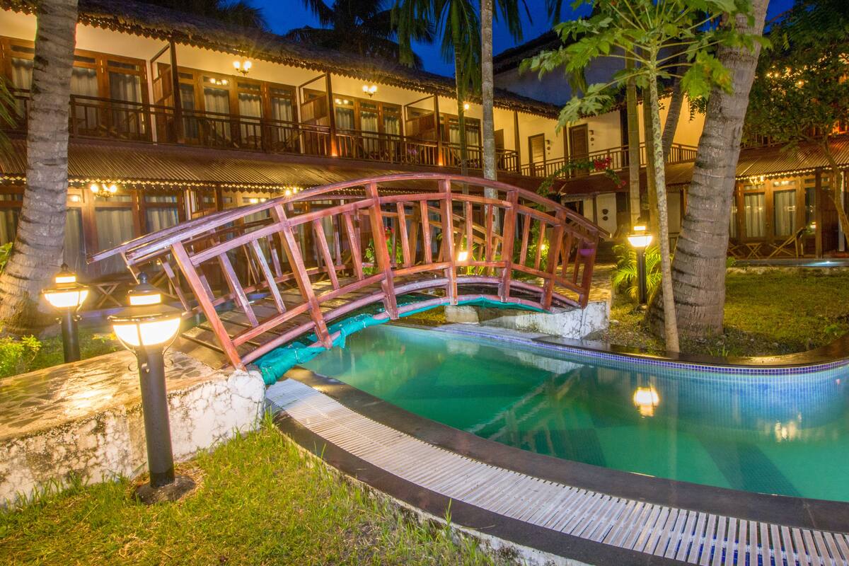 A pool with a wooden bridge, surrounded by palm trees and a building with multiple balconies.