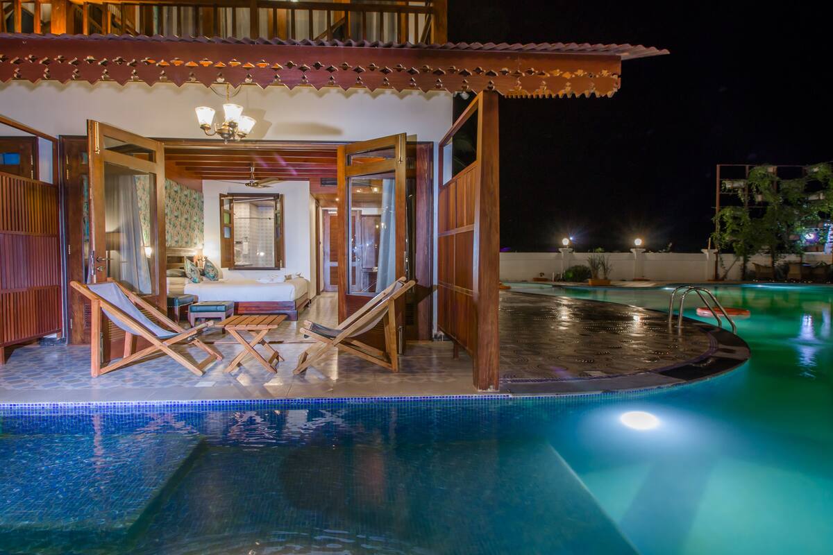A pool area with lounge chairs and a small table, surrounded by wooden structures and a tiled floor.