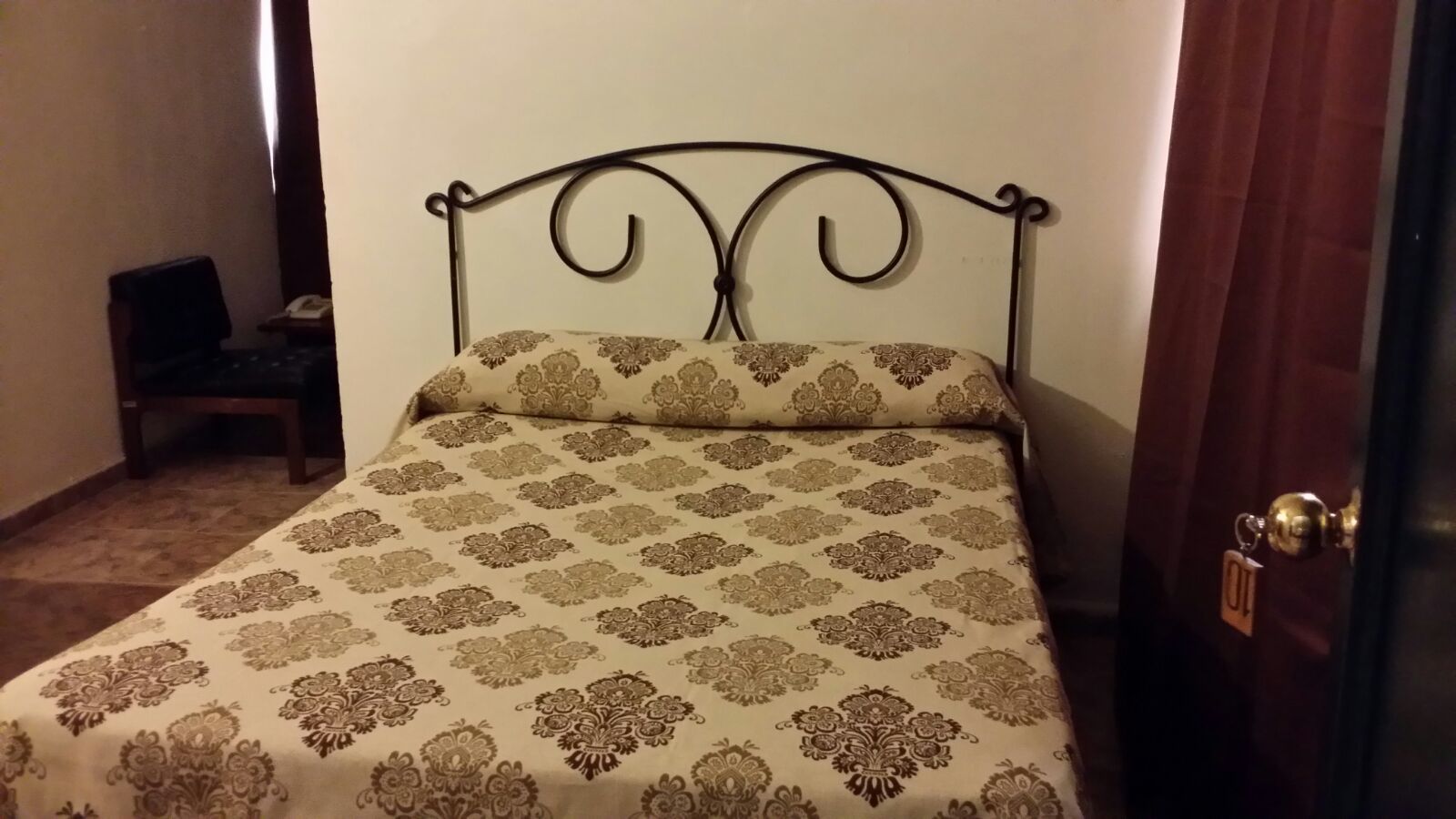 Un lit simple avec une couvre-lit à motifs et une tête de lit en métal, dans une pièce dotée d’une porte et d’une chaise.