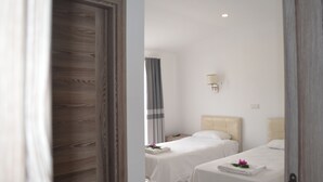 Une chambre d’hôtel avec deux lits, une porte en bois, un rideau gris, une lampe de chevet et une petite table avec une fleur.