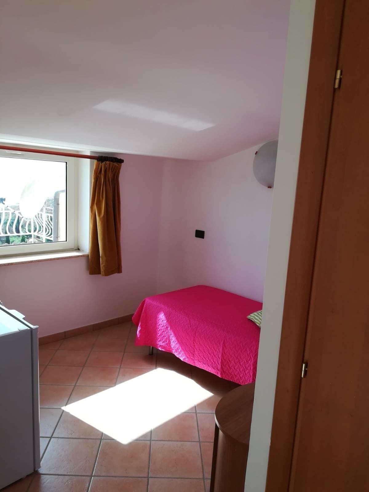 A small, bright room with a bed, a window with a curtain, and a door.