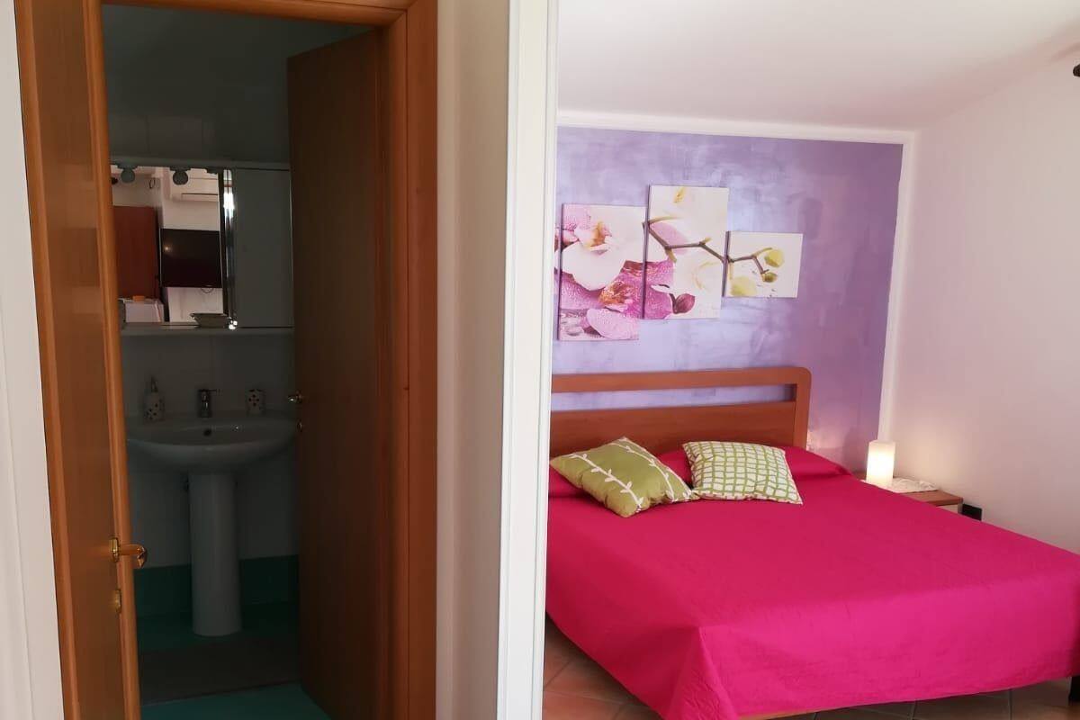 A bedroom with a bed, a nightstand, a wall-mounted shelf, and a bathroom visible through an open door.