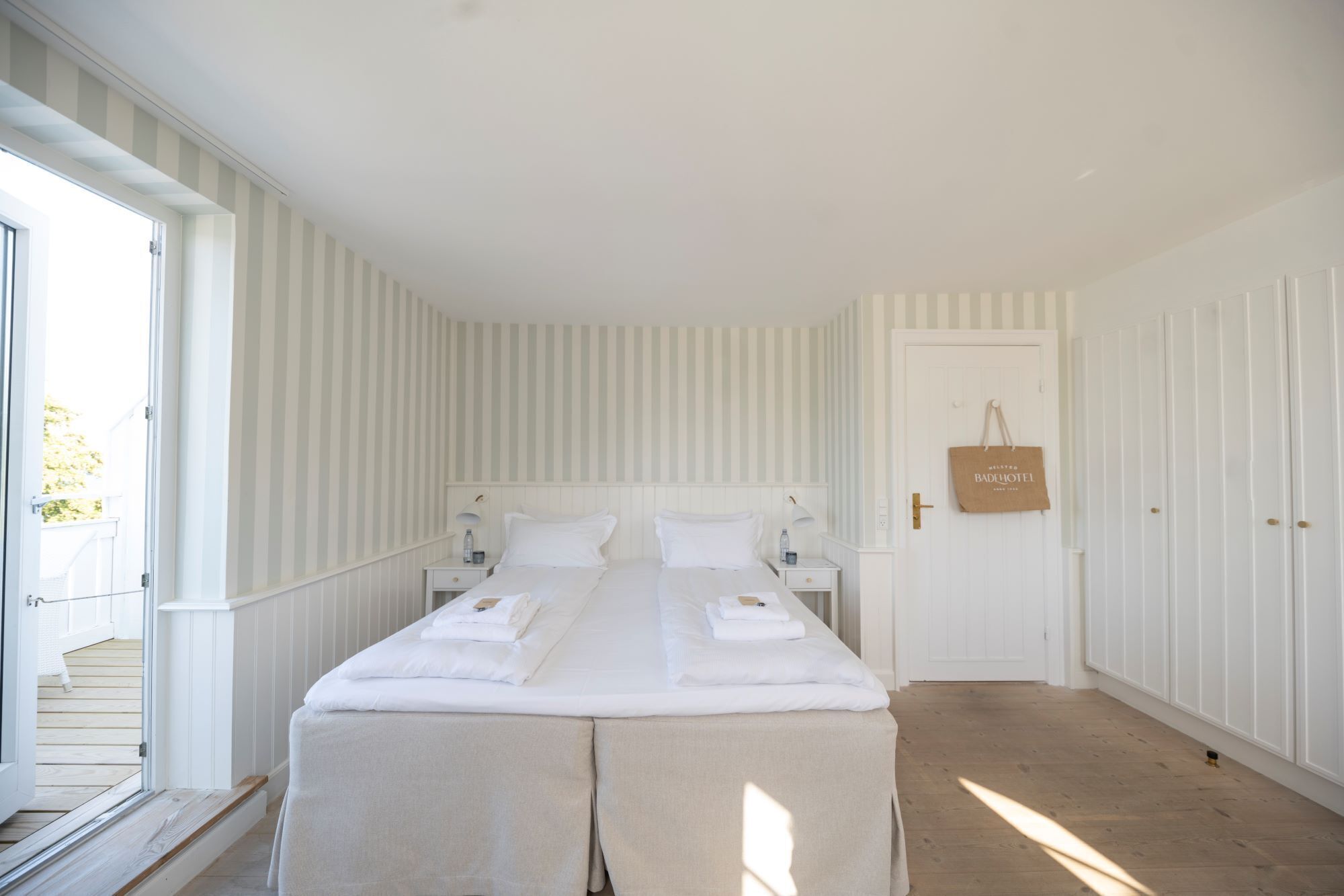 A double bed with white linens, a bedside table with a lamp, a wooden floor, and a striped wall.