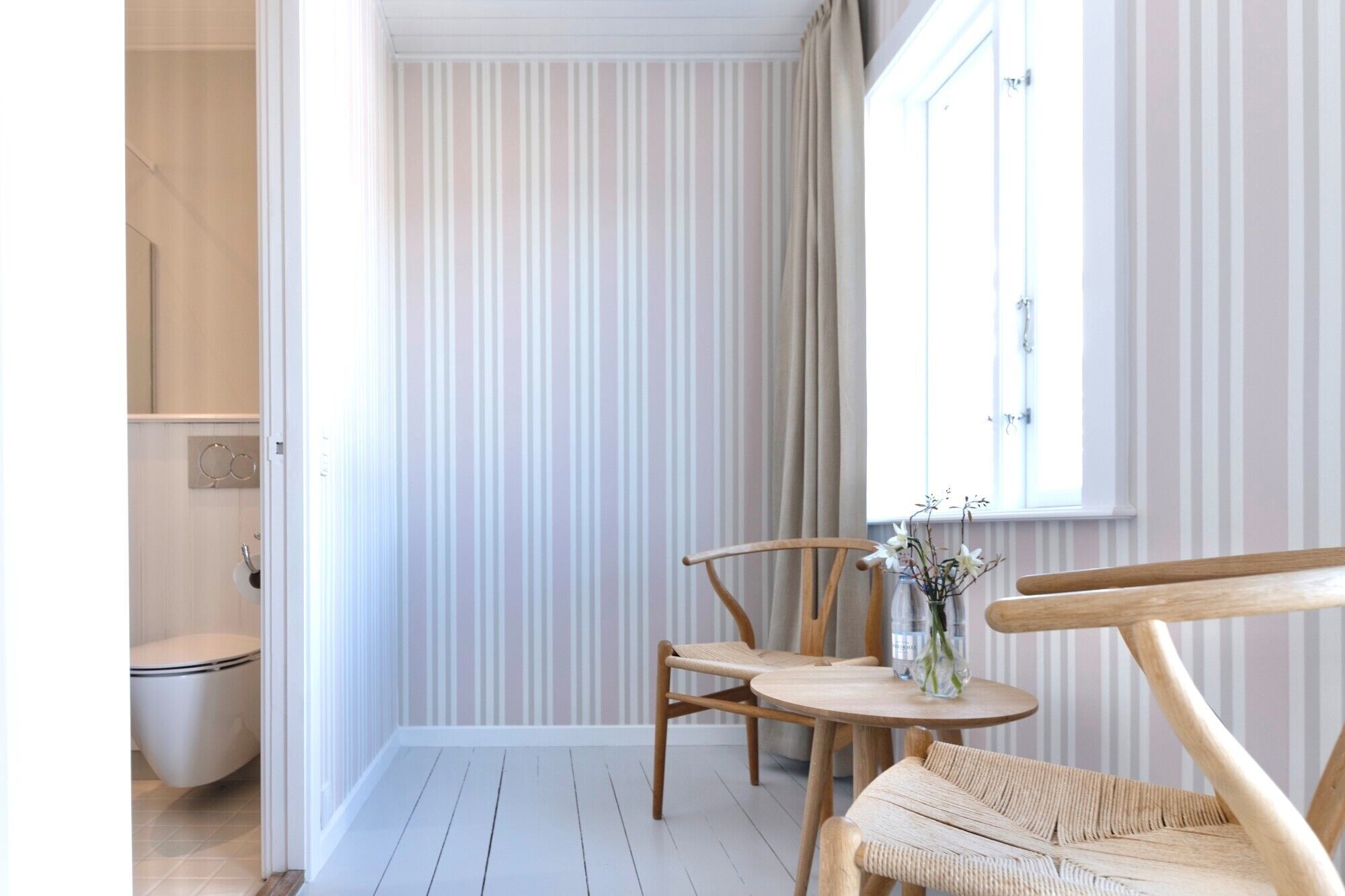 Une salle de bain comprenant des toilettes, un lavabo et un espace douche. Il y a une fenêtre avec des rideaux et une petite table sur laquelle est posé un vase de fleurs.