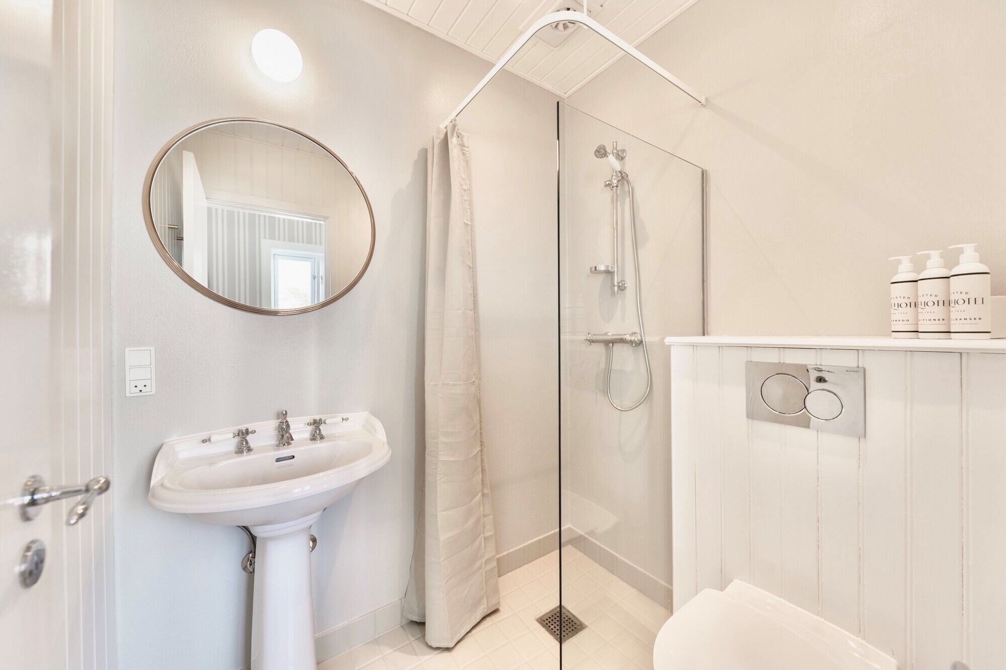 A bathroom with a white sink, mirror, shower, and a vanity with toiletries.
