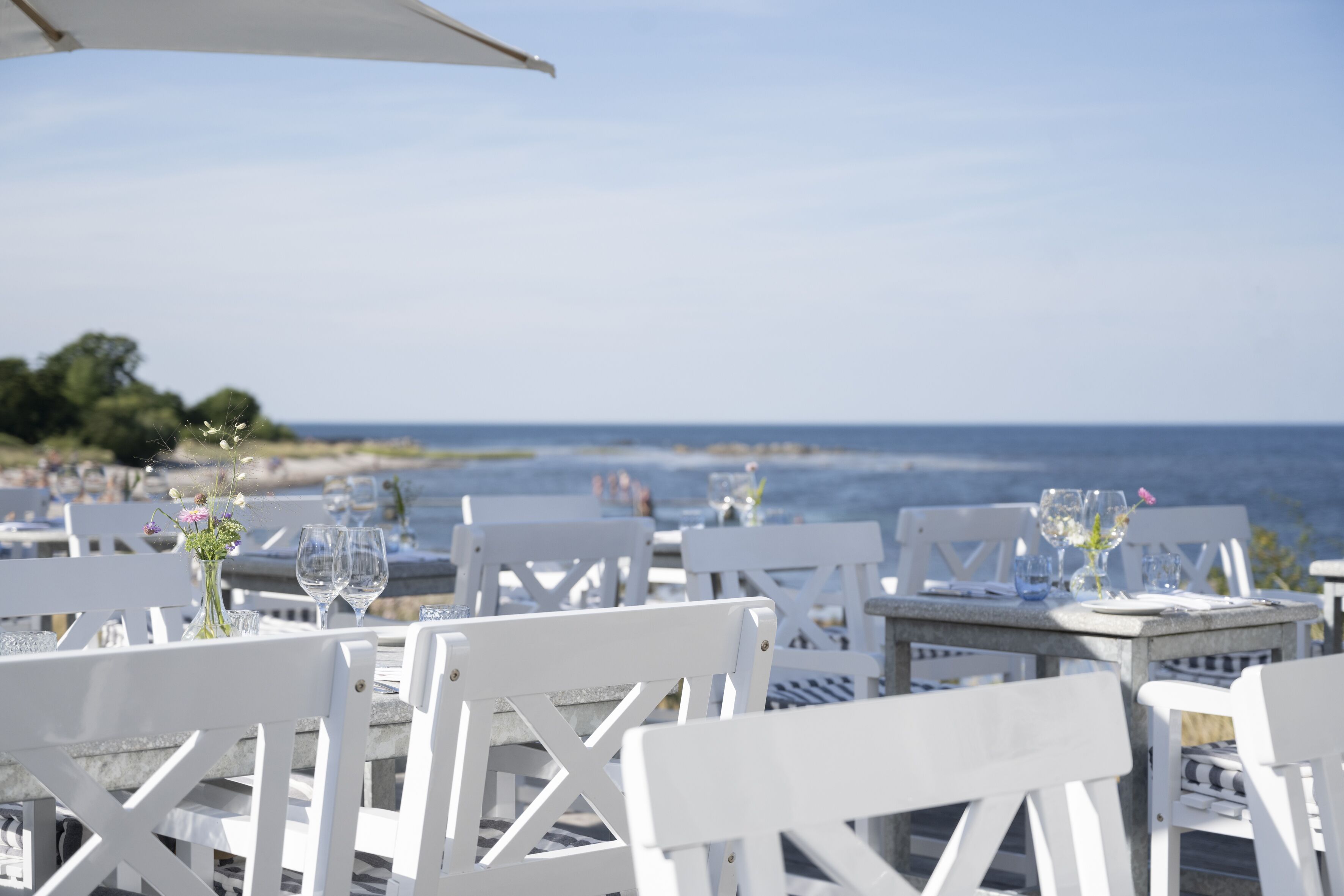 A seaside restaurant with white wooden tables and chairs, set against a backdrop of the ocean and a clear sky.