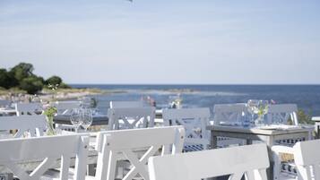 A seaside restaurant with white wooden tables and chairs, set against a backdrop of the ocean and a clear sky.