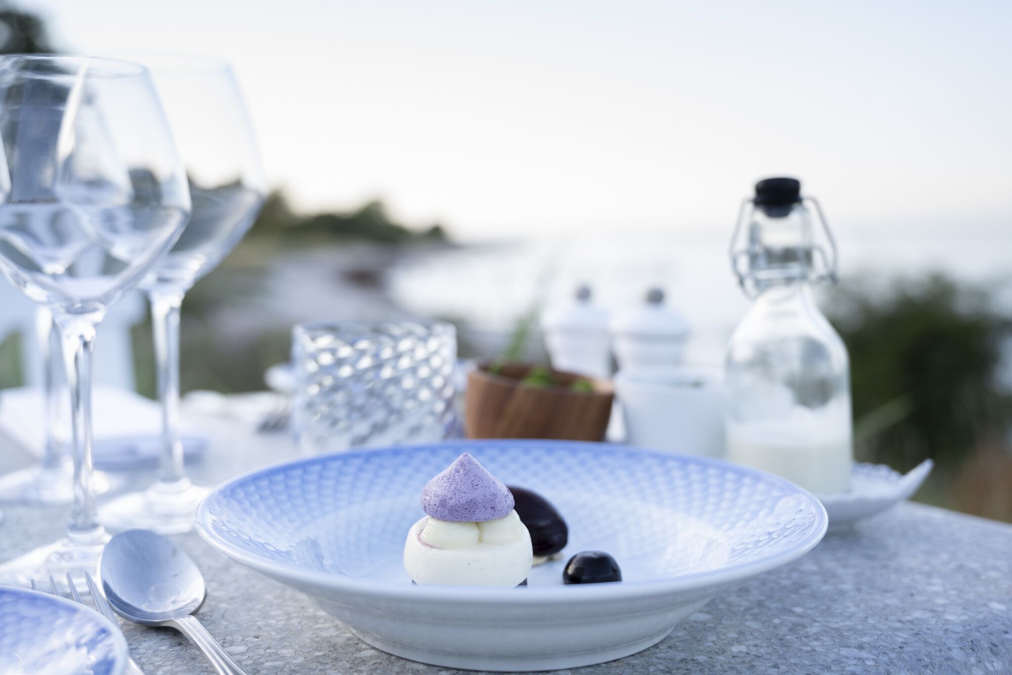 A dessert on a blue plate with a spoon and wine glasses on a table with a blurred background.
