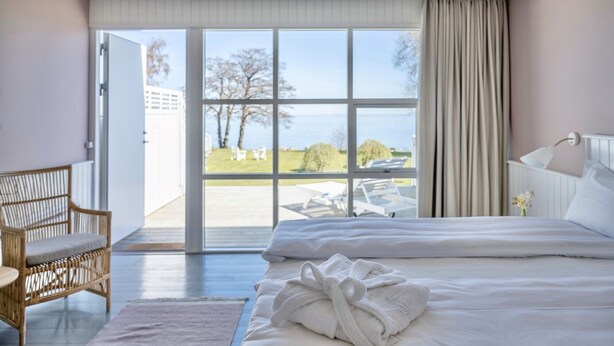 A hotel room with a large bed, a wicker chair, a view of the outdoors through a sliding glass door, and a white bedside table. - Melsted Badehotel  (Gudhjem)