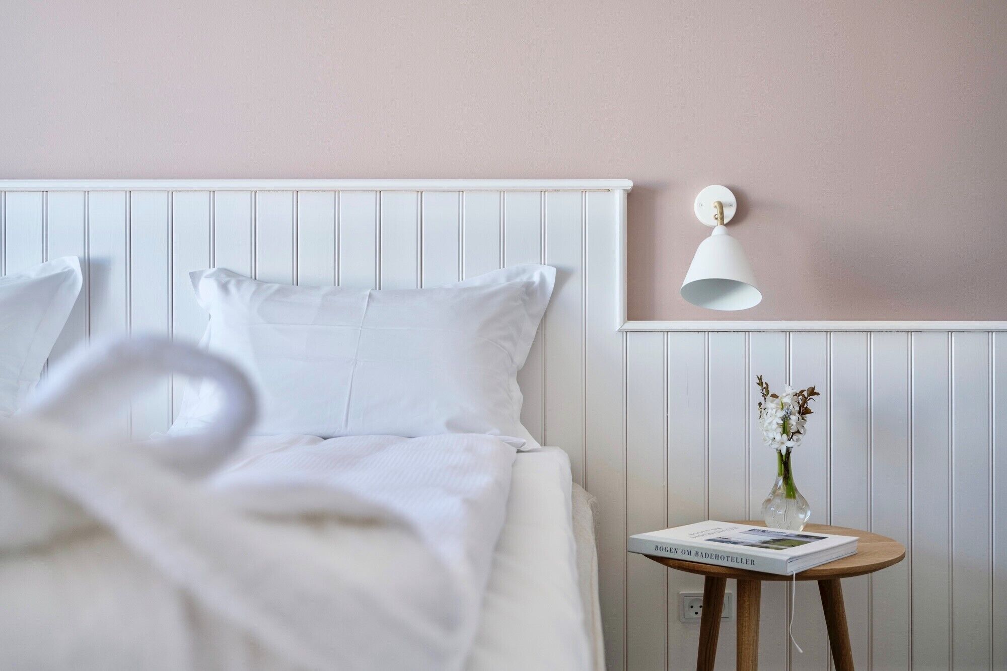 A neatly made bed with white linens and pillows, a bedside table with a book and a vase of flowers, and a wall-mounted lamp.