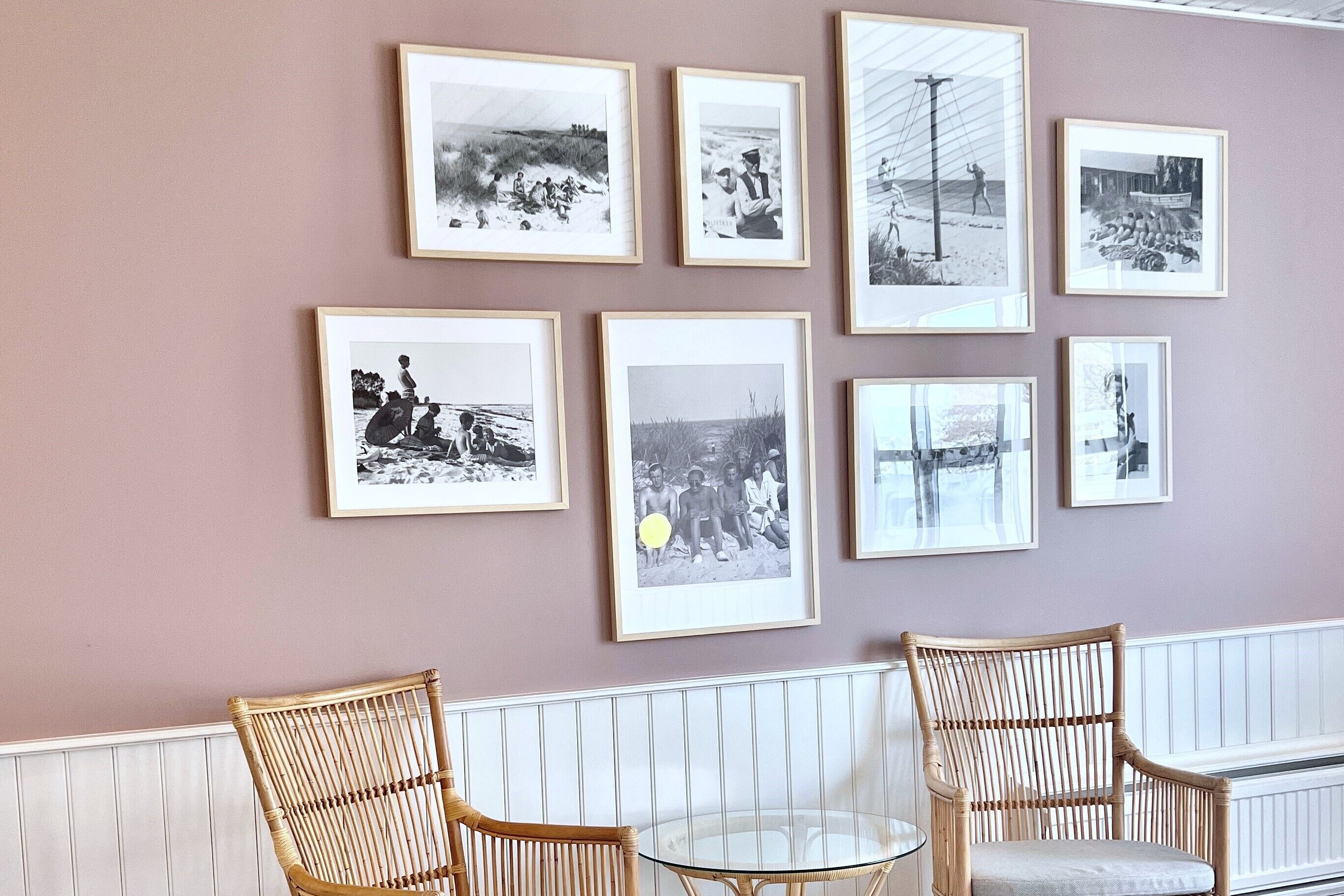 A wall with framed black and white photographs, two wicker chairs, and a glass table.