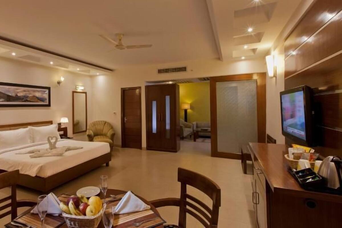 A hotel room with a bed, bedside tables, a TV, a dining area with chairs, and a table set with fruit and a glass.