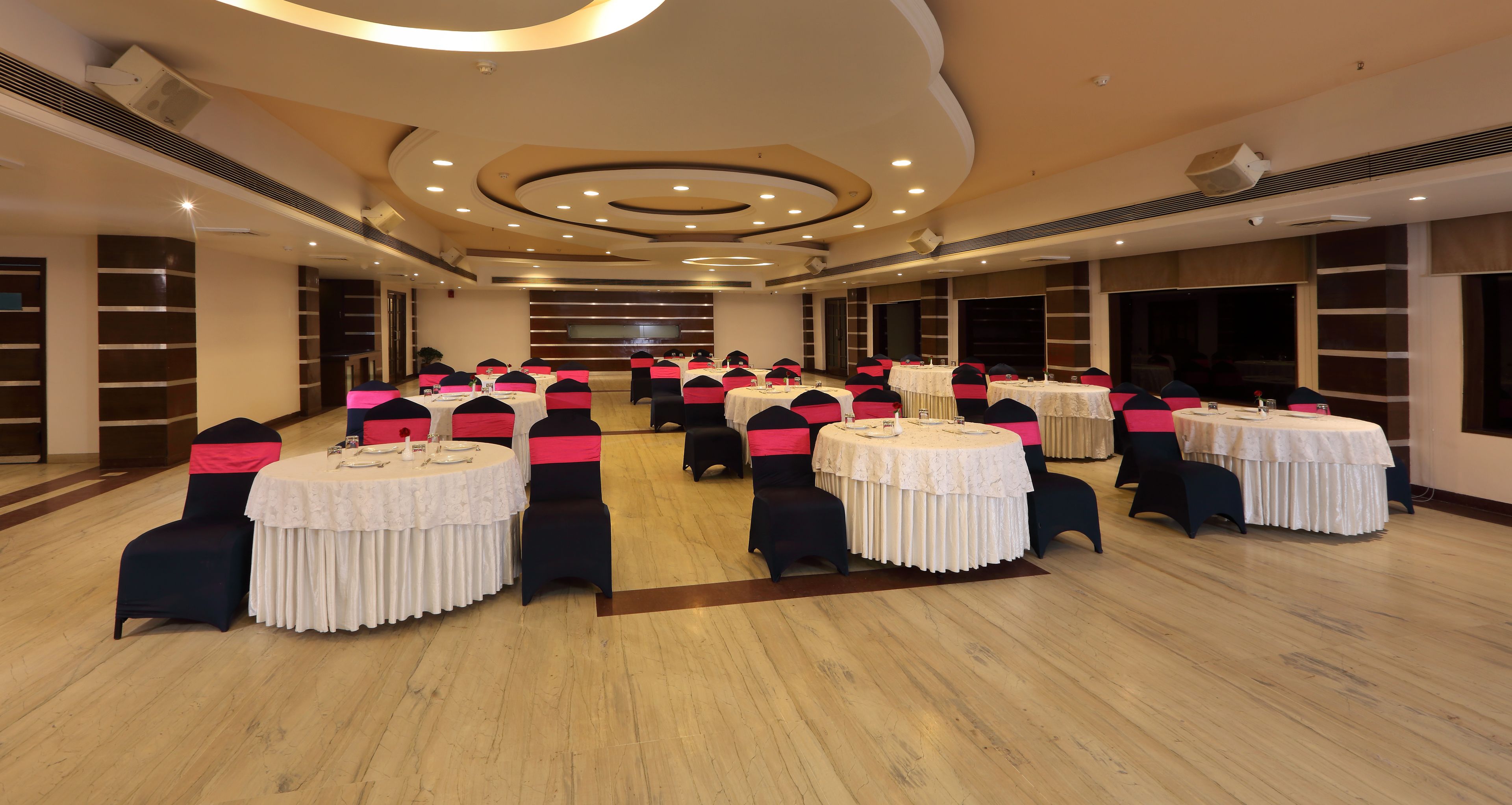 A conference room with multiple round tables, each with a white tablecloth and black chairs with pink sashes. The room has a wooden floor, beige walls, and a modern ceiling design with recessed lighting.