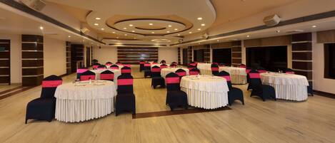 A conference room with multiple round tables, each with a white tablecloth and black chairs with pink sashes. The room has a wooden floor, beige walls, and a modern ceiling design with recessed lighting.
