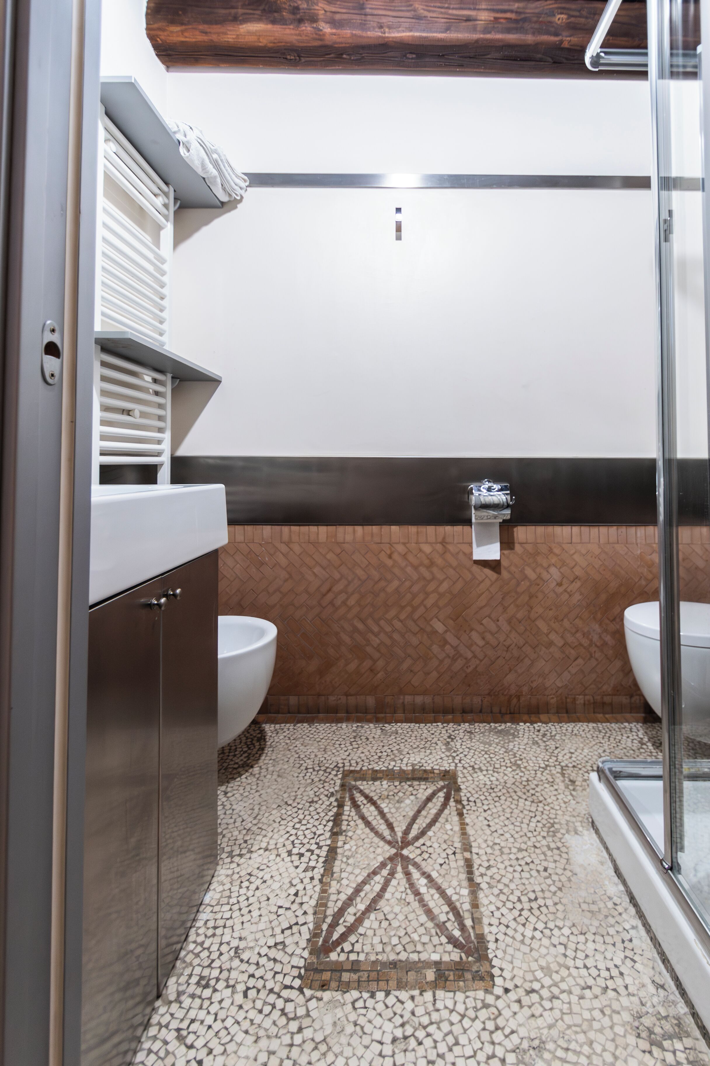 A modern bathroom with a herringbone tile floor, two toilets, a shower area, and a vanity with a towel rack.