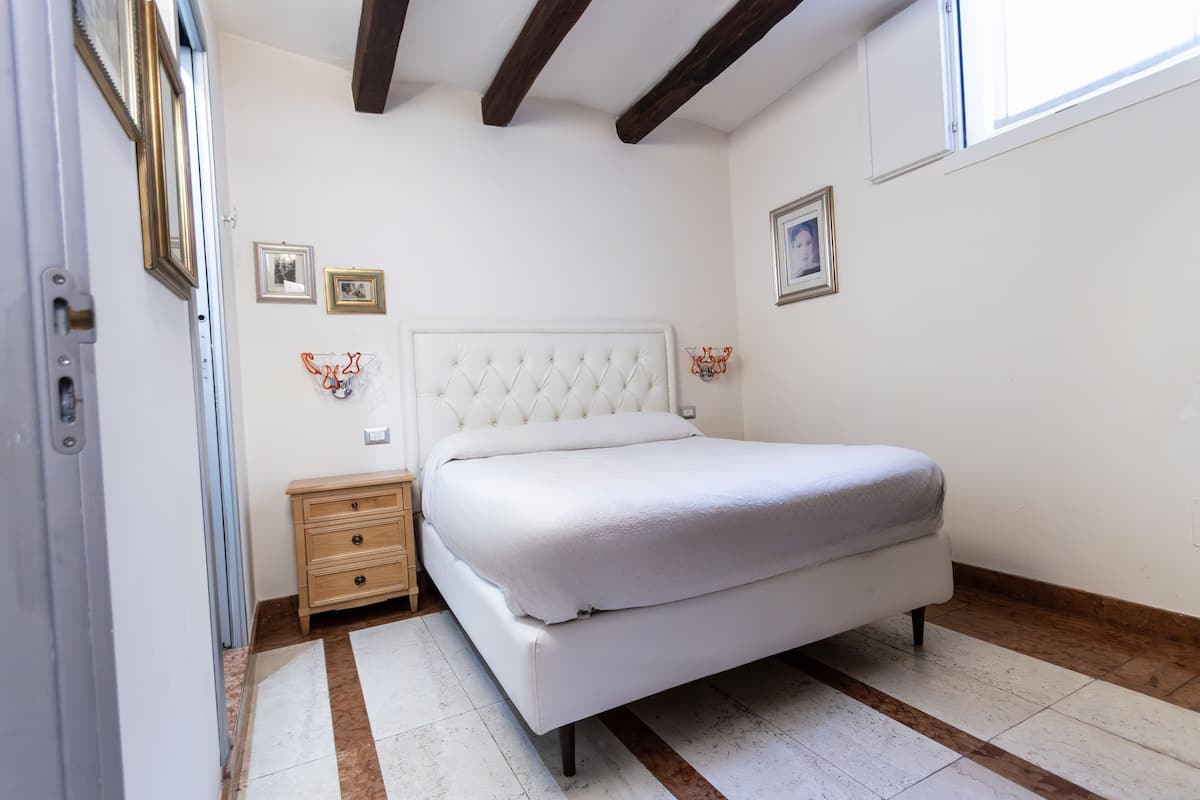 A bedroom with a white bed, wooden nightstand, and a small dresser. There are framed pictures on the wall and a window with a view.