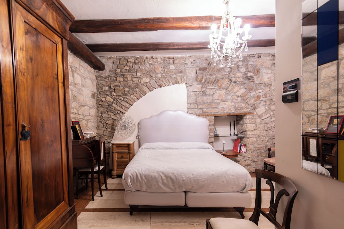 A bedroom with a stone wall, a wooden wardrobe, a bed, a desk, and a chandelier.