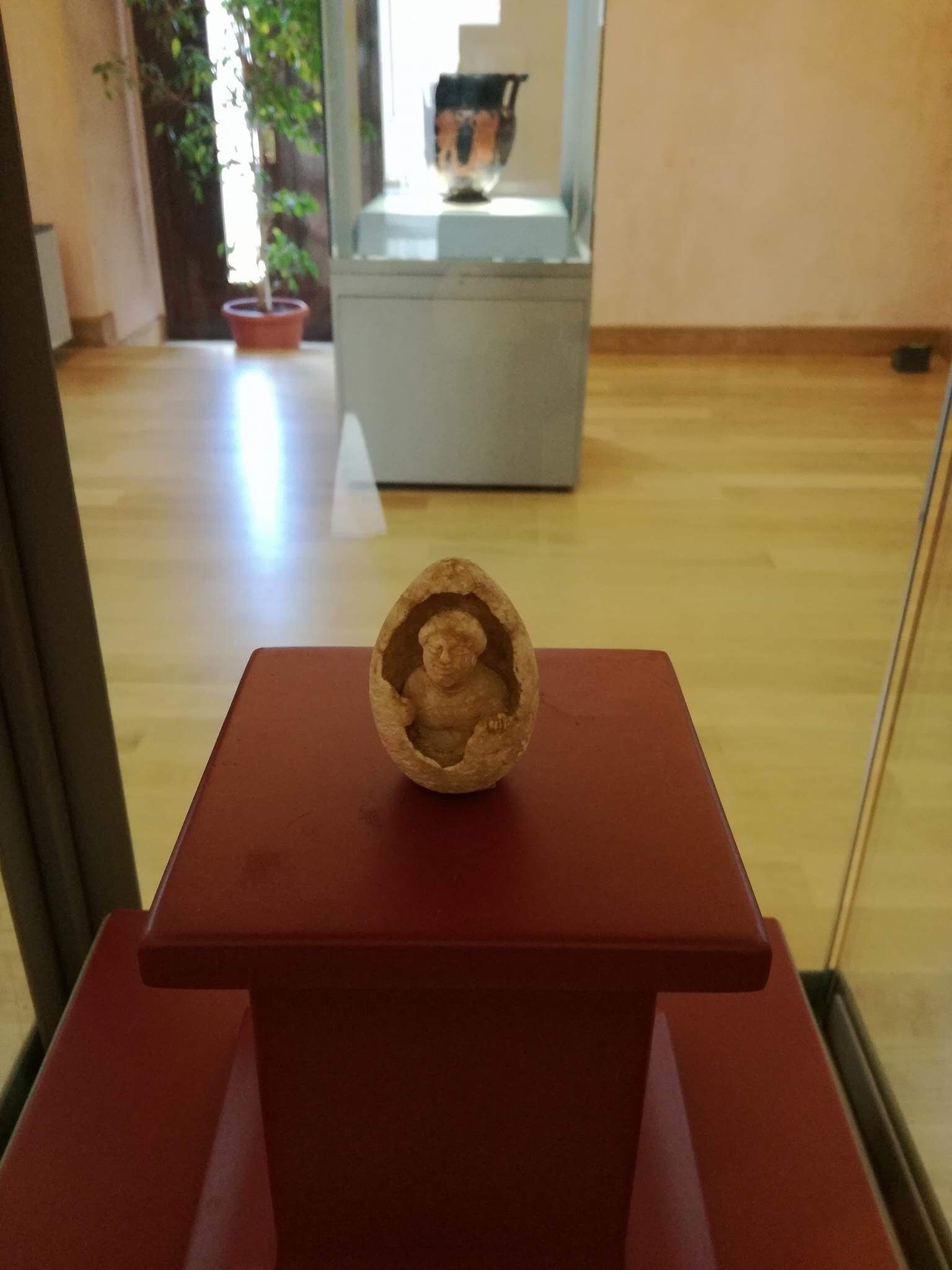 A sculpture on a red pedestal in a room with wooden flooring and a potted plant in the background.