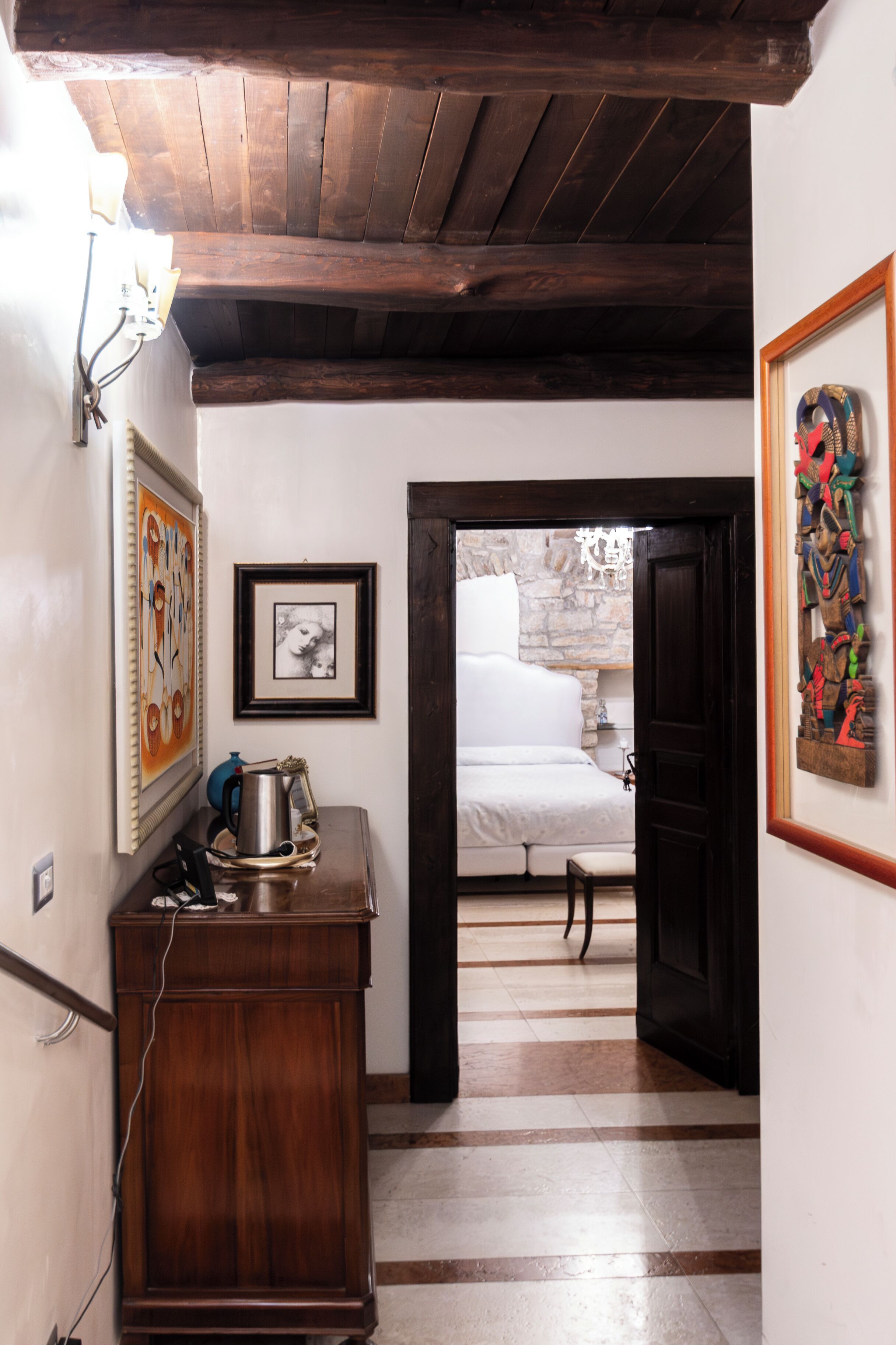 A narrow hallway with a wooden ceiling, a bed in the adjacent room, and a stone wall.