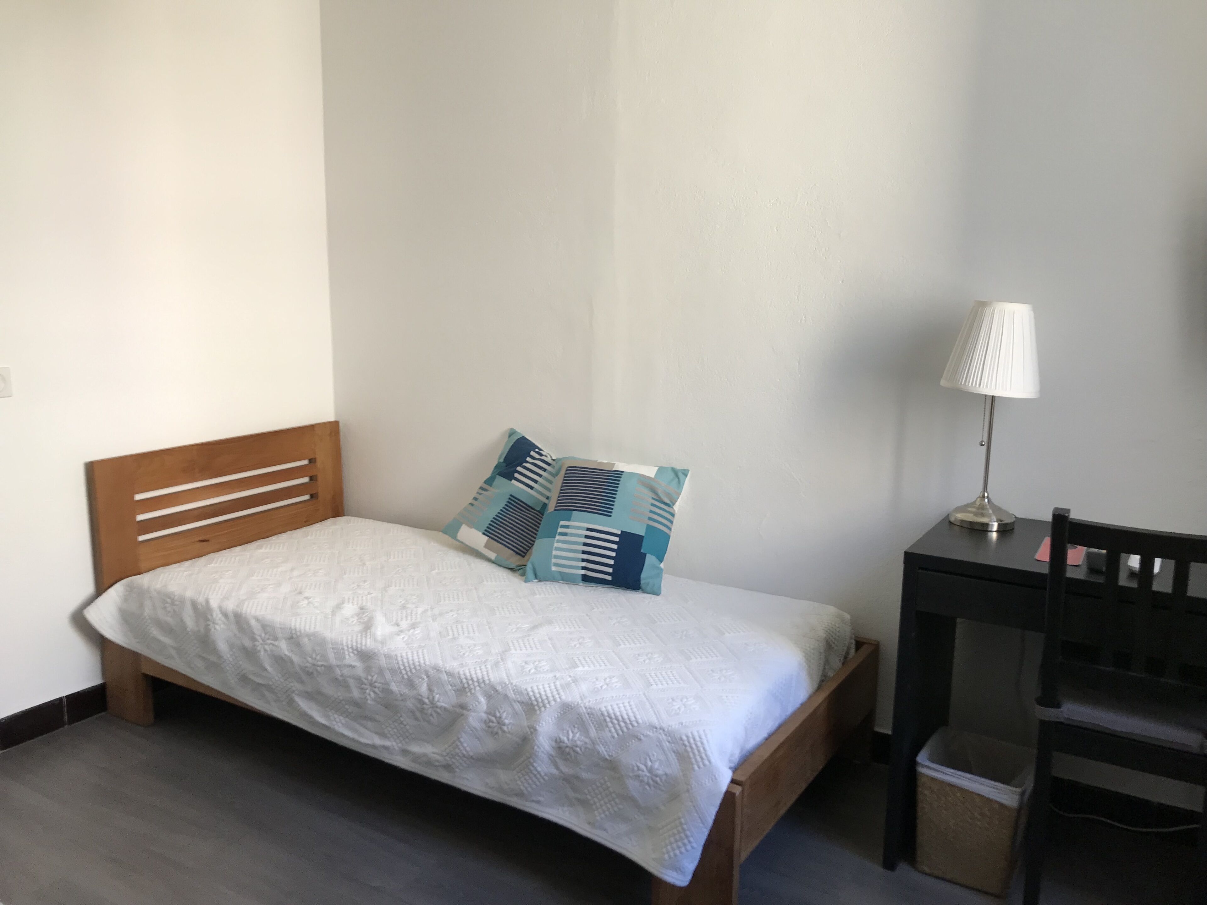 Uma cama de solteiro com cabeceira de madeira, um cobreleite branco e duas almofadas estampadas. Um escritório com luminária e cadeira fica à direita.