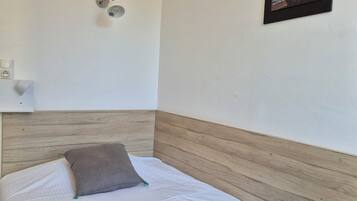 Una cama bien hecha con edredón gris y almohada, cabecera de madera, un enchufe y un cuadro con una imagen del atardecer sobre el océano.