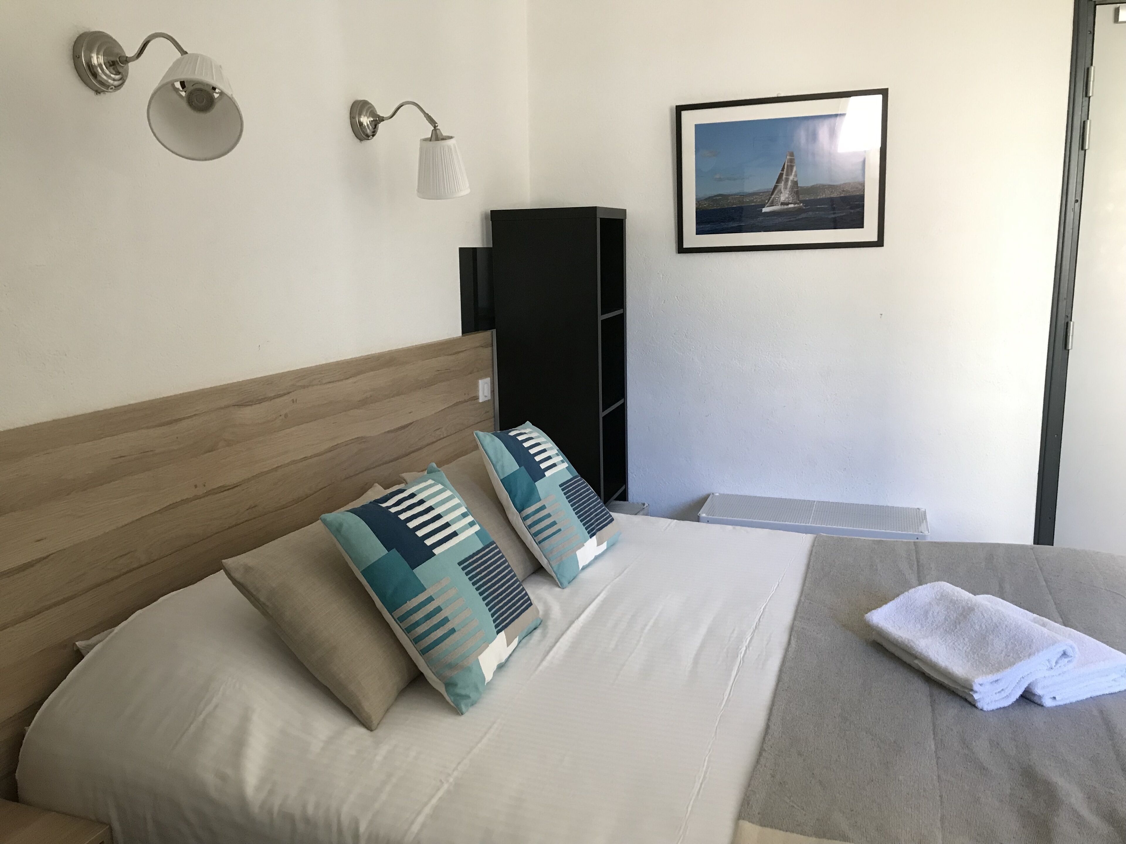 Una cama bien hecha con almohadillas estampadas, una lámpara de noche, un cuadro en la pared y una toalla doblada sobre la cama.