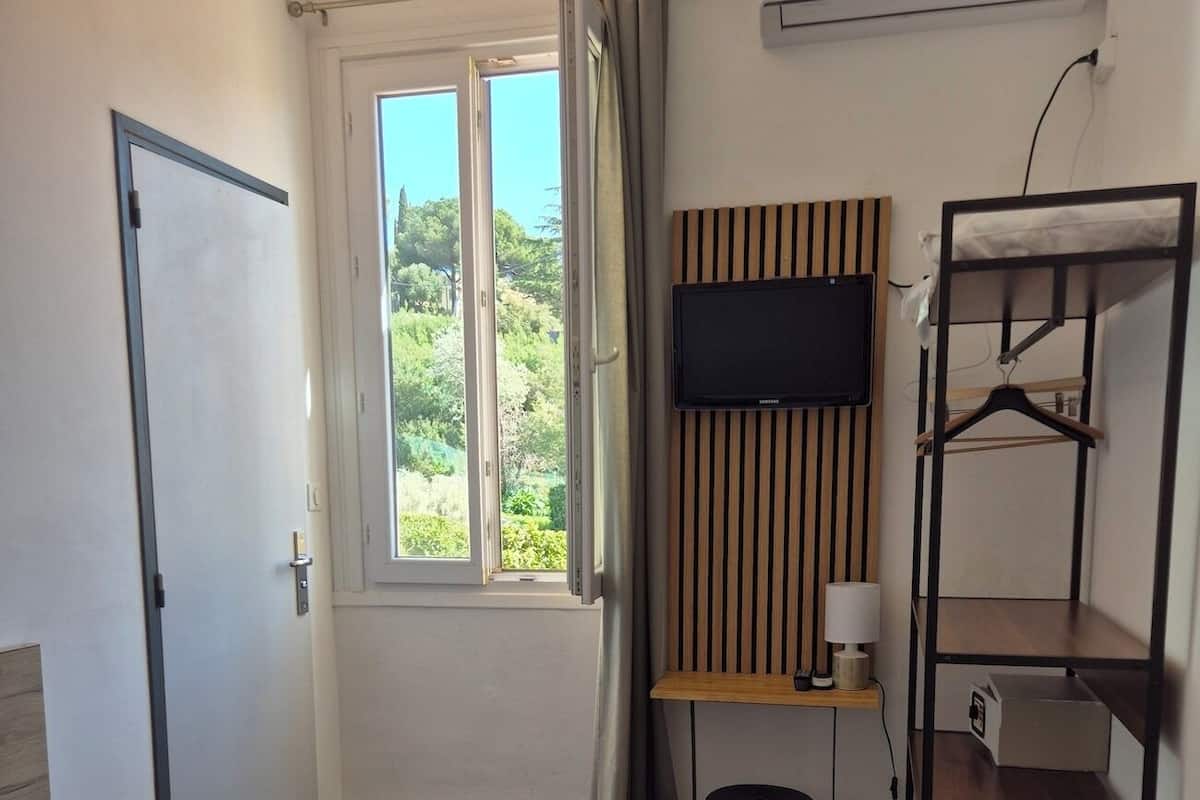 A bedroom with a bed, a TV mounted on a wall, a wooden shelf, and a window with a view of greenery.