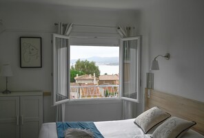 Un dormitorio con cama, ventana con cortinas, lámpara de noche y un cuadro en la pared.