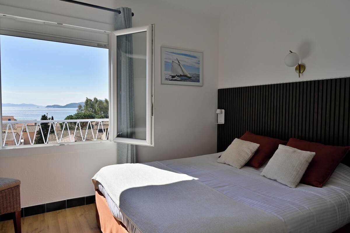 A bedroom with a bed, a window with a view of the sea, and a painting on the wall.