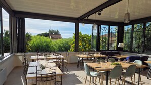 Un comedor al aire libre iluminado por el sol, con mesas y sillas, ventanas amplias y vistas a zonas verdes y edificios.