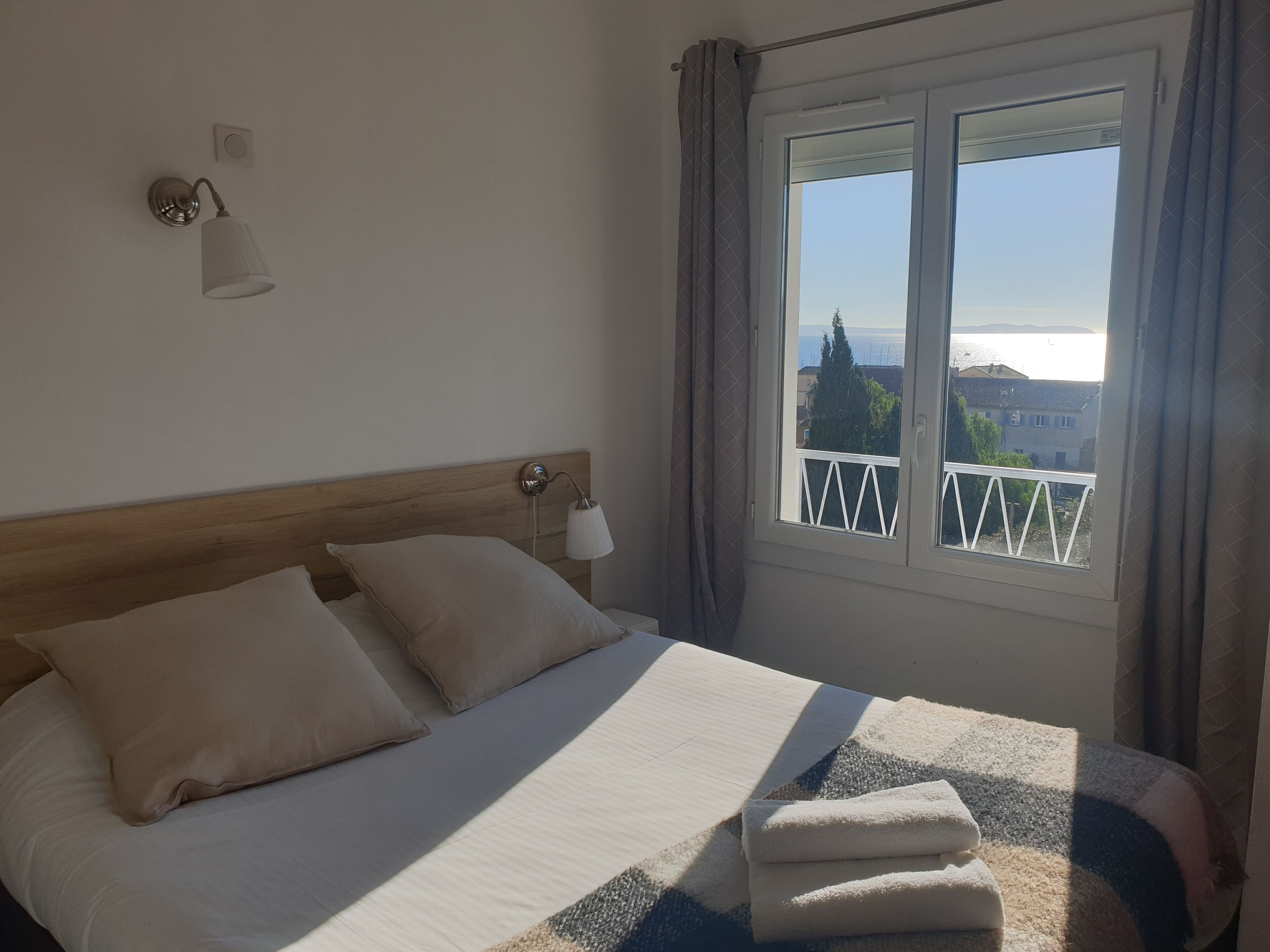 A hotel room with a bed, a nightstand, a lamp, a window with curtains, and a view of the sea and buildings.