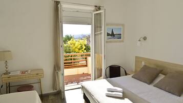 A hotel room with a bed, a desk with a lamp, a chair, and a view of the outdoors through an open door.