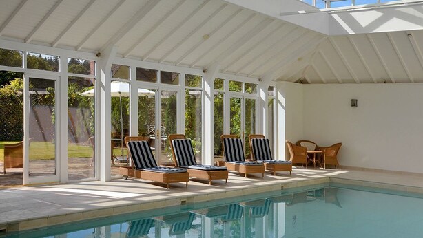 Ein überdachter Poolbereich mit Liegestühlen und einem Tisch, umgeben von großen Fenstern.