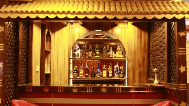 Una zona de bar con mostrador de madera, estantes llenos de botellas de licores y taburetes rojos.